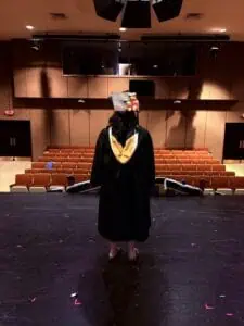 Student with long dark hair standing on a dark theatre stage in a black graduation gown with her back to the camera. The theatre is lit in the back with rows of empty red seats. Her graduation cap has colorful decorations on top and her stole has gold trim.