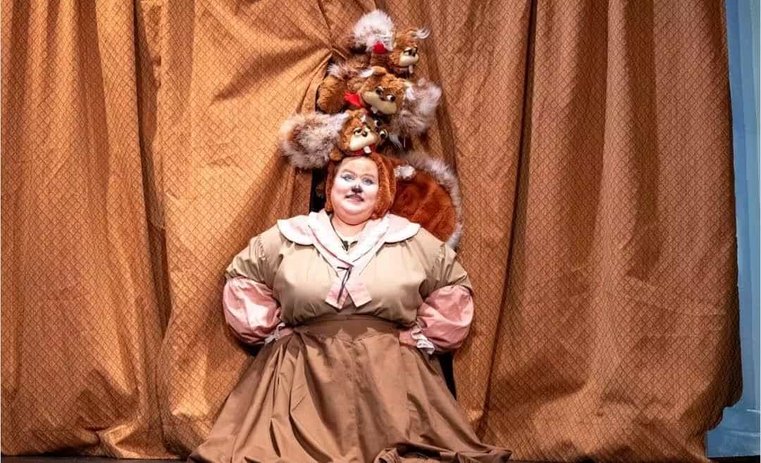 Student in costume as squirrel with bushy tale and an old-fashioned dress on, with four brown squirrel puppets over her head, in front of a gold-orange theatre curtain