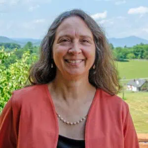 Inge Terrill stands outside on a sunny day with green trees and grassy fields, mountains, and small house behind her, wearing a dark dress, pearl necklace, and a coral cardigan 