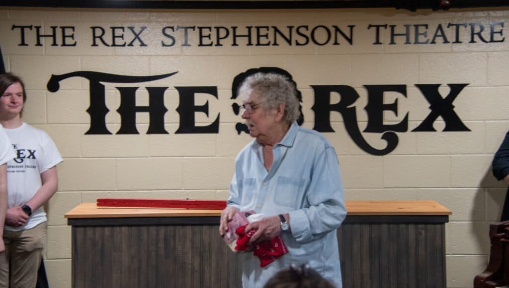Rex Stephenson Theatre lobby with Rex standing right in front of the silhouette of his head on the wall. He has white hair and a light blue shirt. A student in a white Rex Stephenson Theatre shirt is standing at the left edge. The red divining rod is on the box office counter behind Rex.