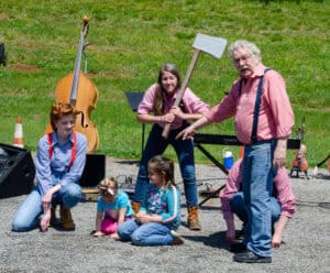 Three Jack Tale Players in red and blue gingham shirts and suspenders in front of musical instruments outdoors, with one holding a fake axe and Rex Stephenson pointing to two small girls in a Jack Tale show