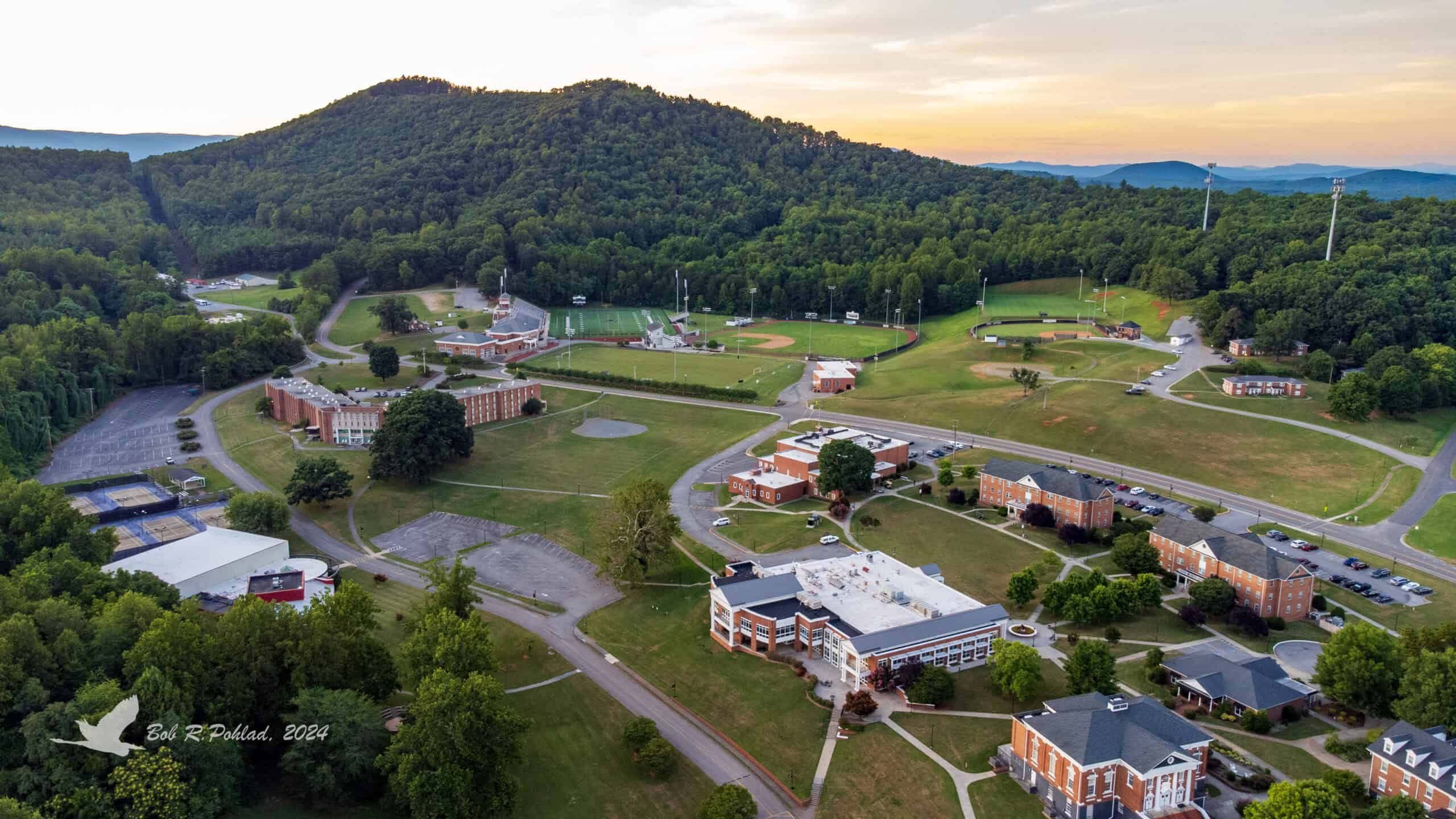 Aerial view of Ferrum college with multiple brick buildings, athletic fields, and parking lots, set against a backdrop of forested hills at sunset.