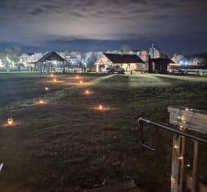 Lighted pavilion at farm museum with lights of campus in the distance and small lanterns across the field