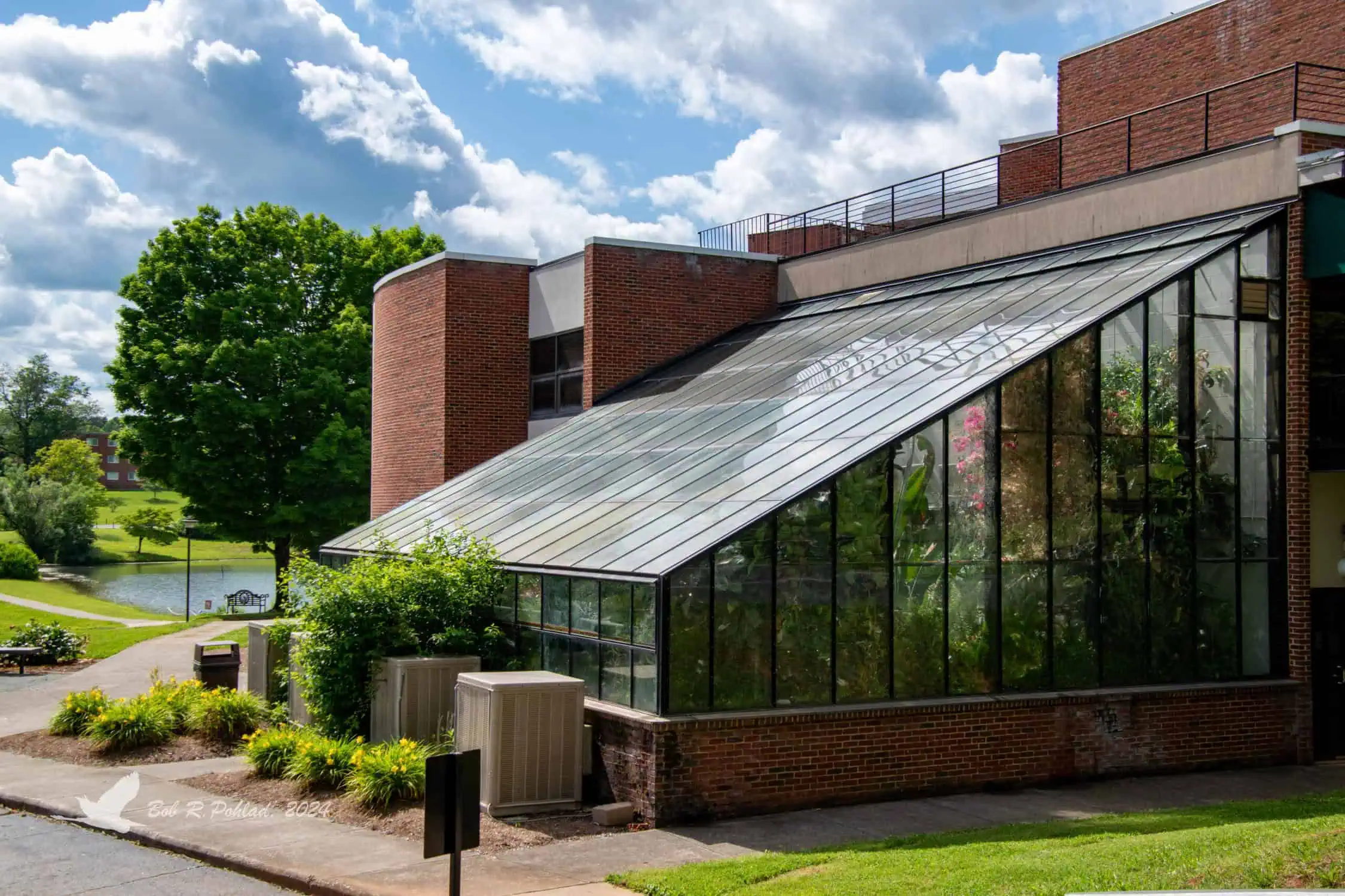 Garber Hall greenhouse with slanted glass roof attached to brick science building, with greenery outside and sky of puffy clouds
