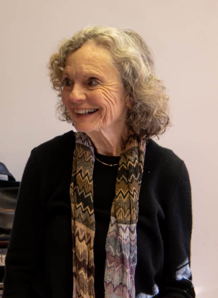 Fair Robey standing against a light wall in a dark outfit with a long straight scarf in grey and russet muted tones
