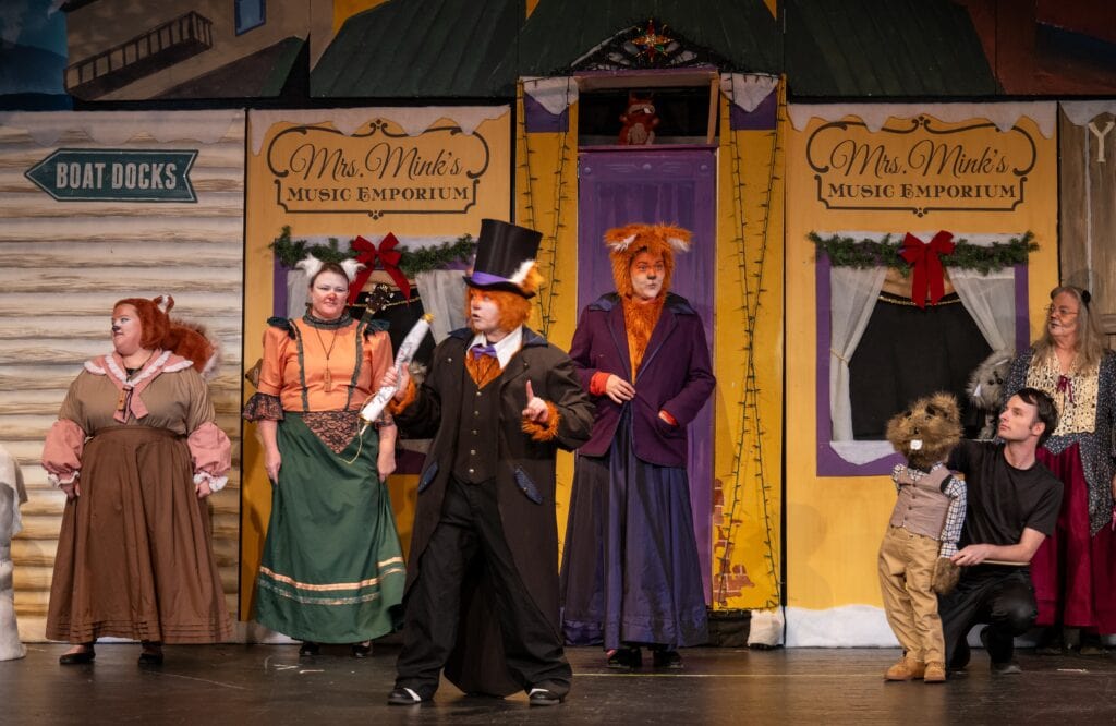 Five female actors standing in animal costumes and vintage clothing in front of a set of store fronts, with several animal puppets around them and one male puppeteer in black with a large woodchuck puppet.