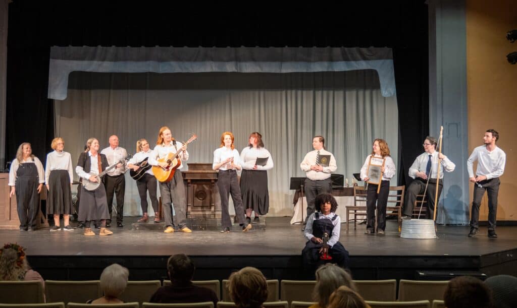 A long row of performers of different ages on a black stage in white shirts and black skirts or pants, with plain curtains behind them. Some hold instruments. A young one sits on the edge of the stage and some audience members' heads can be seen dimly in the foreground. A large antique pulpit can be seen in the middle behind the actors.
