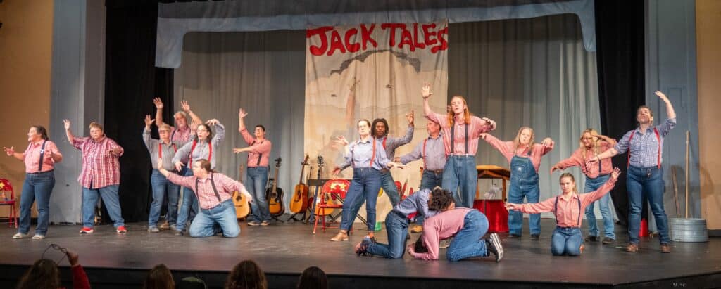 Jack Tale Players in red and blue gingham shirts and jeans perform in a lively scene on stage with old Jack Tales banner as backdrop