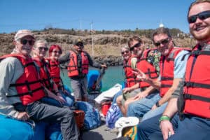 Group of travelers in orange life vests in two rows on small boat in water with Galapagos shore behind them