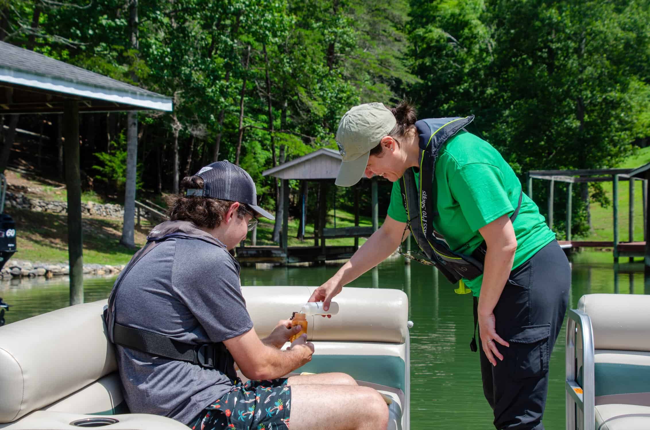Photo of faculty member in bright green t-shirt and ball cap bending over and smiling while assisting student dressed in grey summer clothes sitting on boat doing scientific sampling, with lake landscape in background