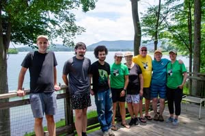 One row of students, faculty, staff, and community members in front of a railing in summer clothes, some with logo of Smith Mountain Lake Water Quality Program on their shirts, with lake and trees behind them