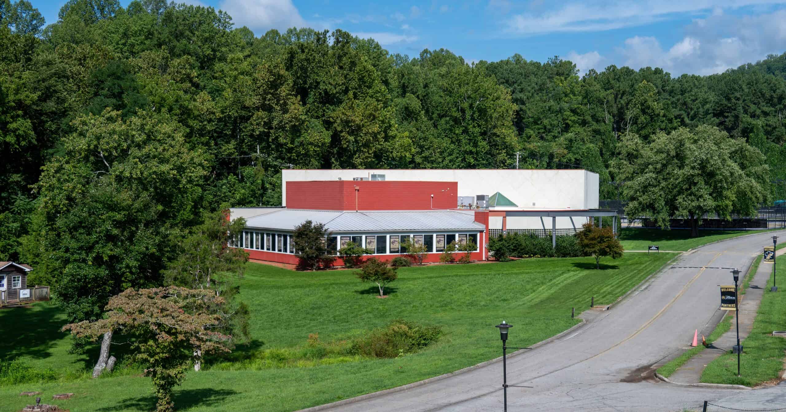 Fitness Center building surrounded by grass and tall trees. A red and white modern low building with windows on lower left and half of front.
