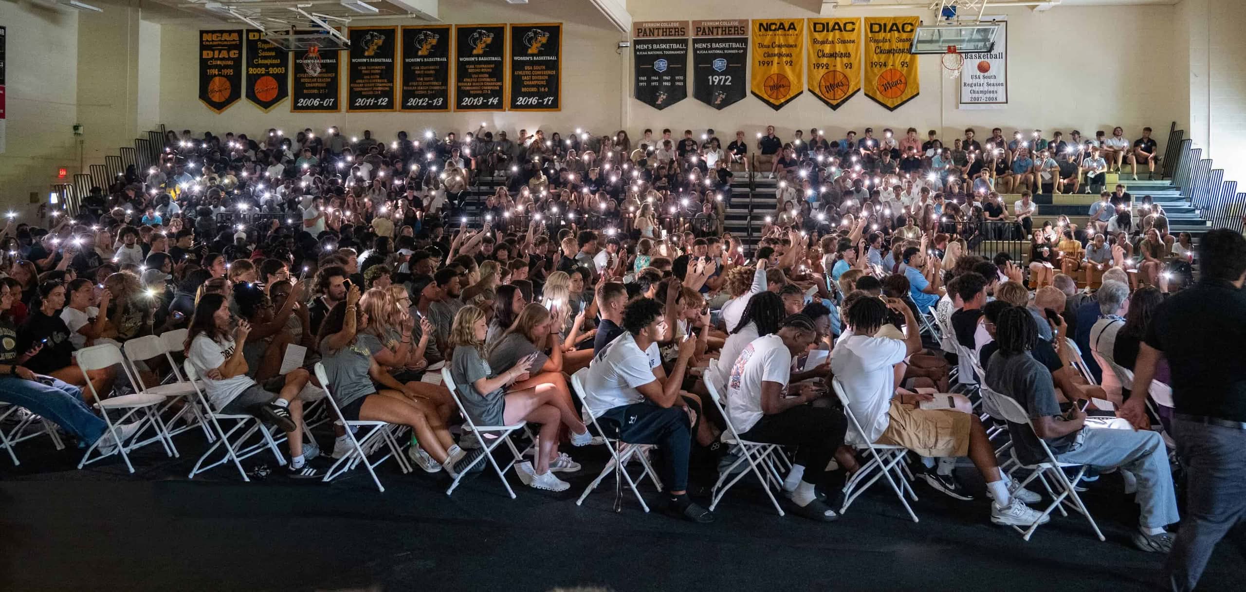 Gym filled with crowd of students with their phone lights on, sports banners on the back wall.