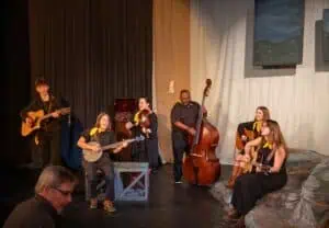 Orchestra Appalachia, 6 players in black clothes and gold kerchiefs, playing in the Rex Stephenson Theatre, some sitting down with their instruments, with dark and light curtains behind them and part of a painting of mountains showing. They have guitars, banjo, bass, and fiddle.