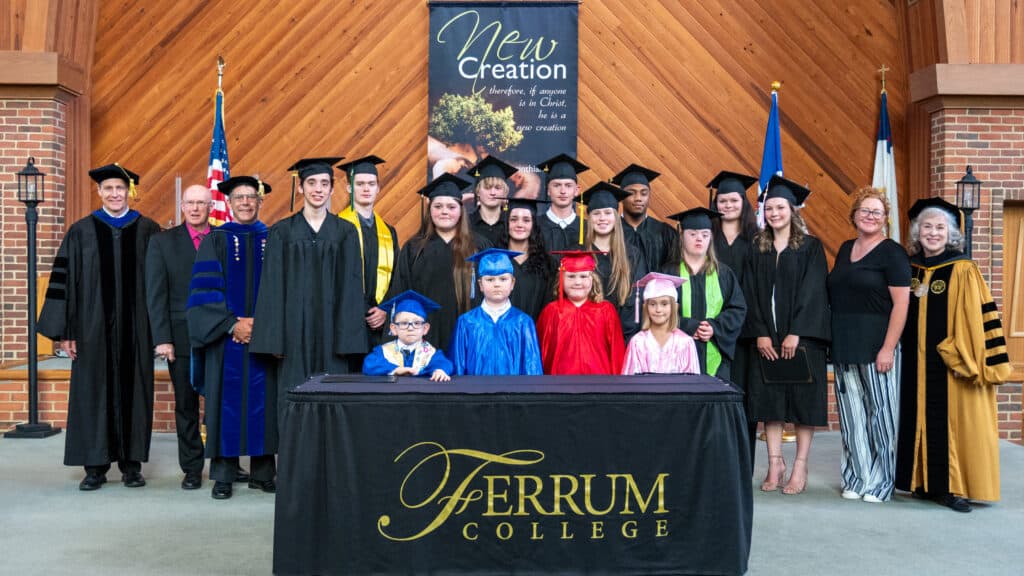 A group of homeschool graduates pose after there graduation ceremony flanked by Ferrum College officials with flags and banners in the background