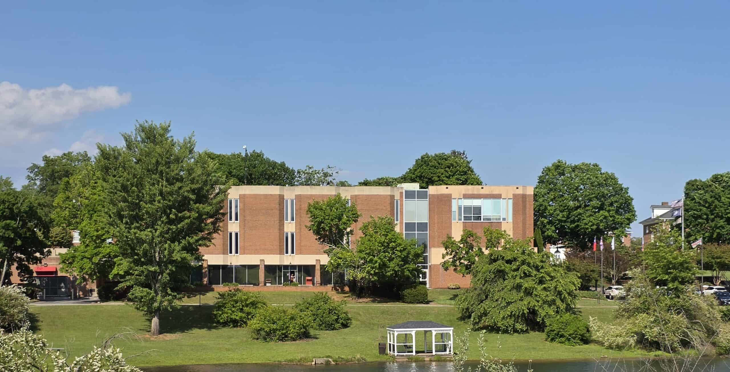 Ferrum College campus with small lake in foreground with gazebo and bushes, modern brick library and trees in background
