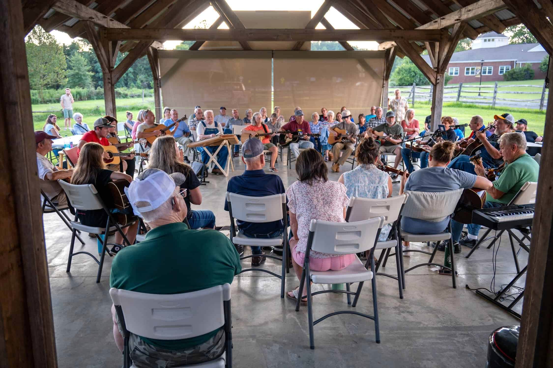 Wooden farm pavilion crowded with seated musicians in the center and listeners all around them, wearing summer clothes with bits of farm field behind the pavilion.