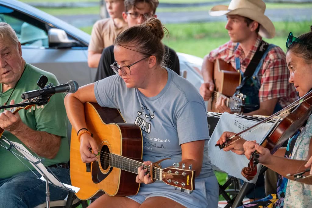 Student in glasses, grey t-shirt and shorts, with dark hair on top of her head, plays guitar while professor playing fiddle looks on from the right, surrounded by other musicians with a car and green grass behind them.