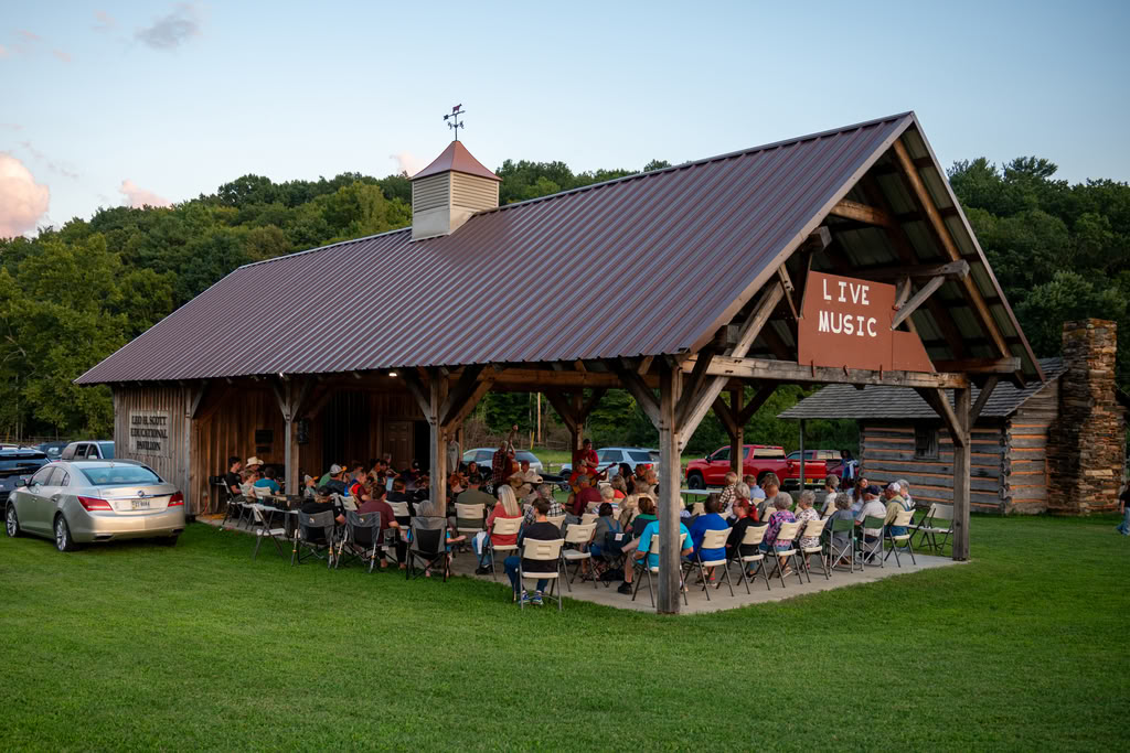 Wooden farm pavilion crowded with seated musicians in the center and listeners all around them, wearing summer clothes with