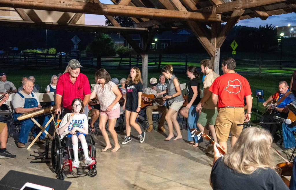 Inside wooden farm pavilion in evening shadows, a line of dancers in summer clothes follow Bobby in red shirt pushing a woman in wheelchair in inner circle, with musicians and listeners behind them.