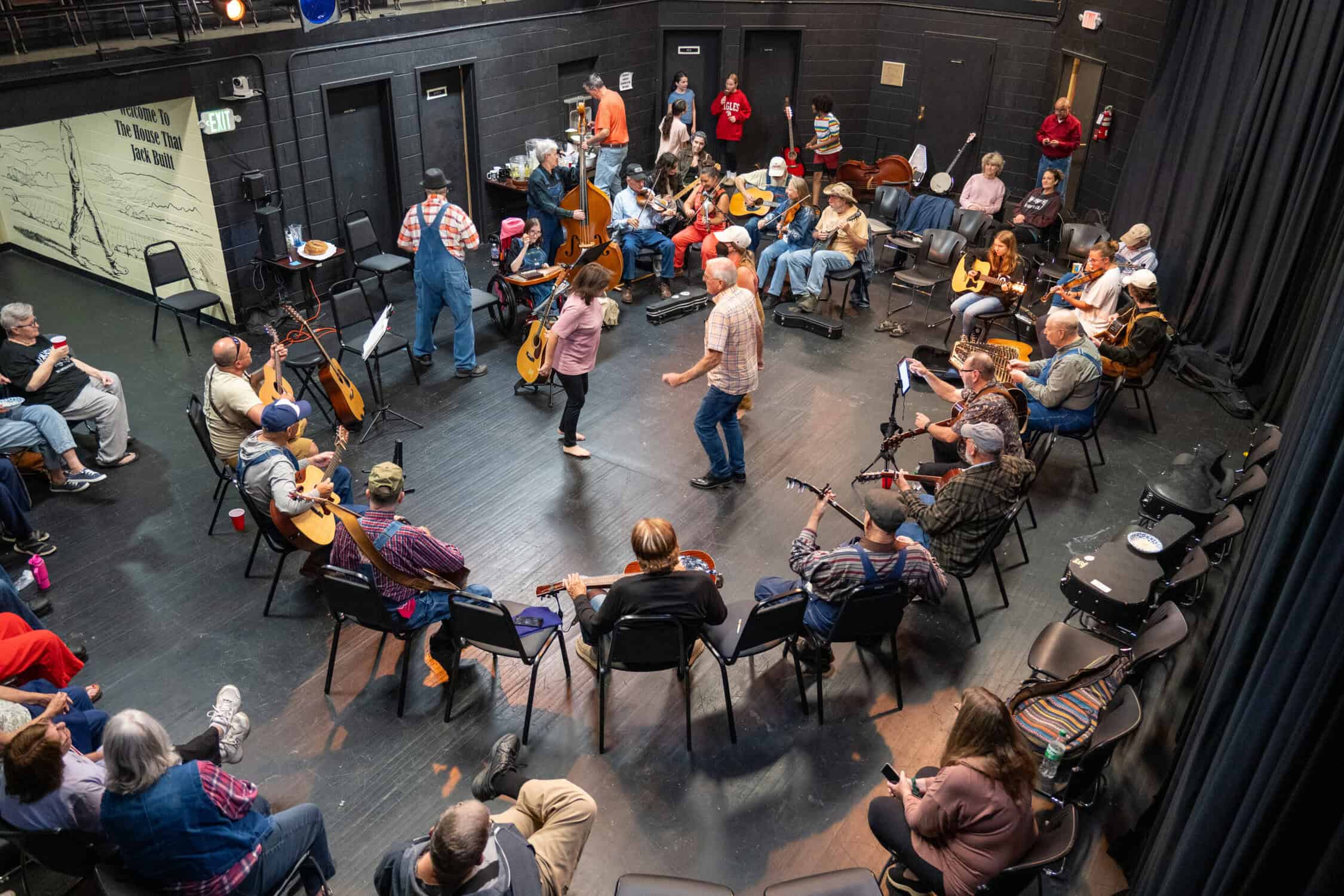 Crowd of people of all ages in a black theatre below a mezzanine, playing traditional instruments in a circle, with people dancing in the middle, and listeners around the outside circle, and a food table in back