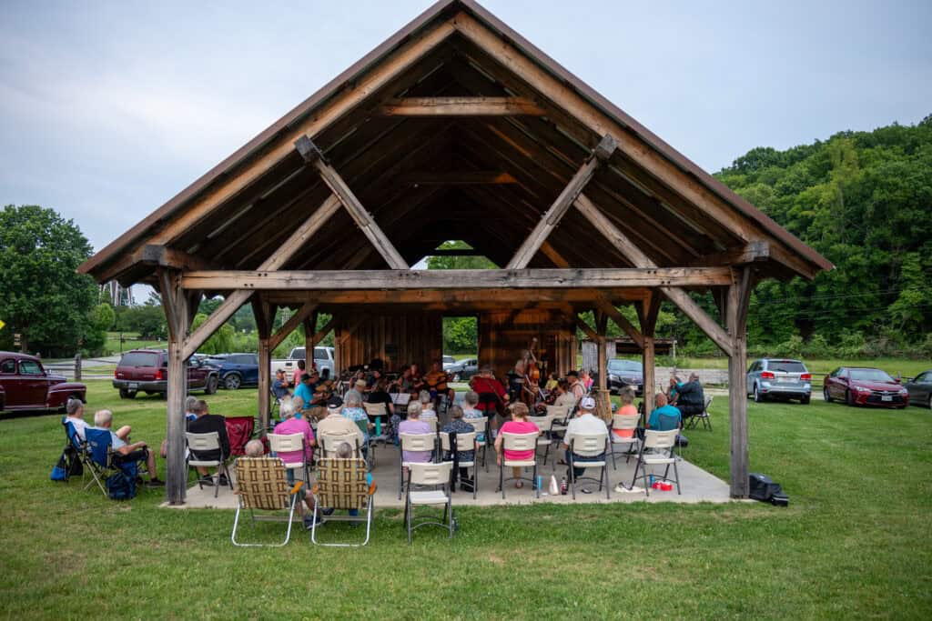 wooden pavilion with pointed, beamed roof, surrounded by grass and green hills, crowded with musicians and listeners sitting at music jam