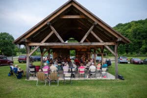 wooden pavilion with pointed, beamed roof, surrounded by grass and green hills, crowded with musicians and listeners sitting at music jam