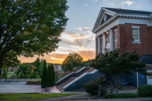 Schoolfield Hall classical red brick and white building from an angle toward front of building with large front steps, patio at bottom, sunset and trees in background