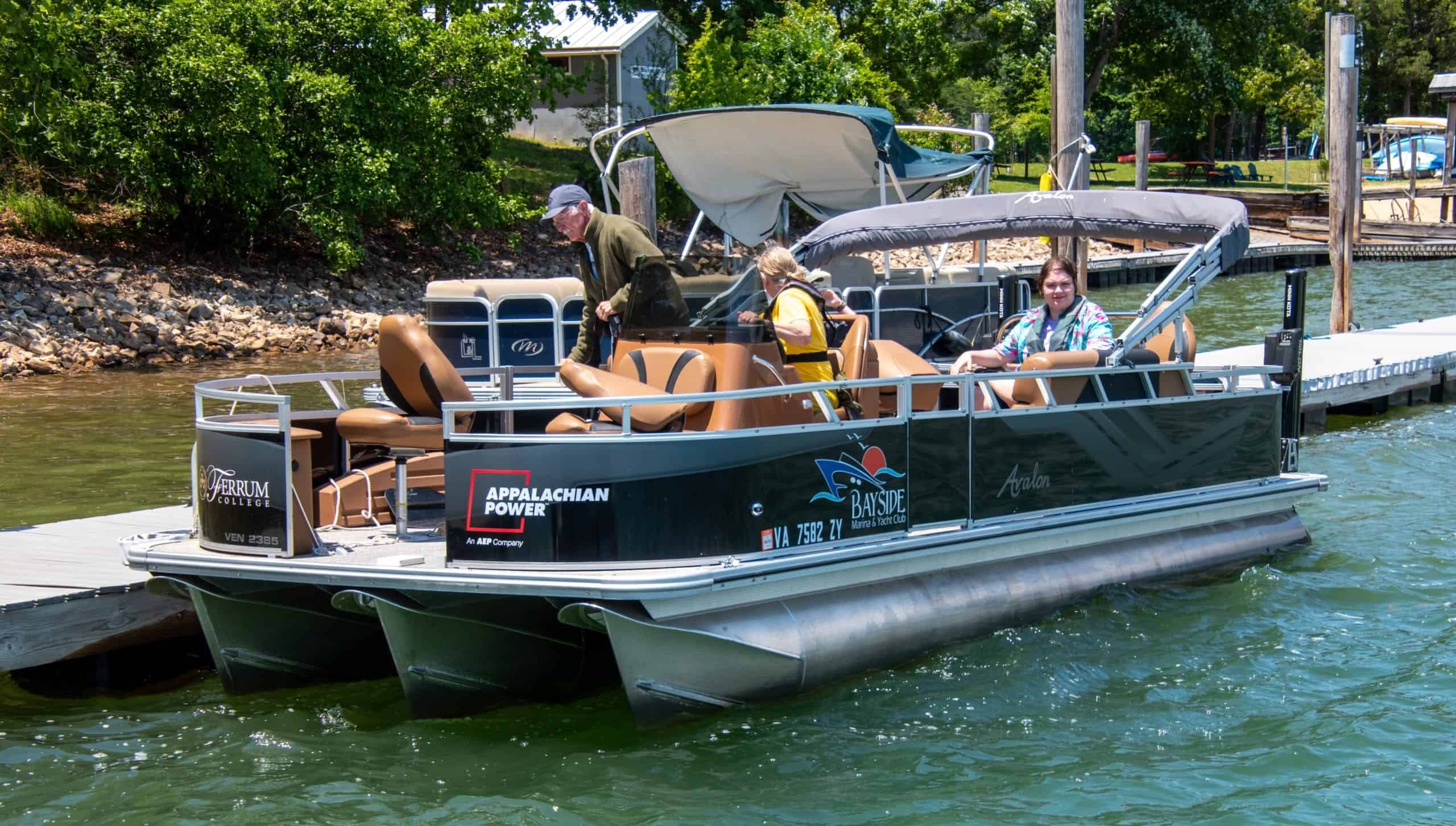 Scientists and students in summer clothes on water quality black motor boat with grey awning, tan seats, and white trim, near lake shore with buildings and trees in back