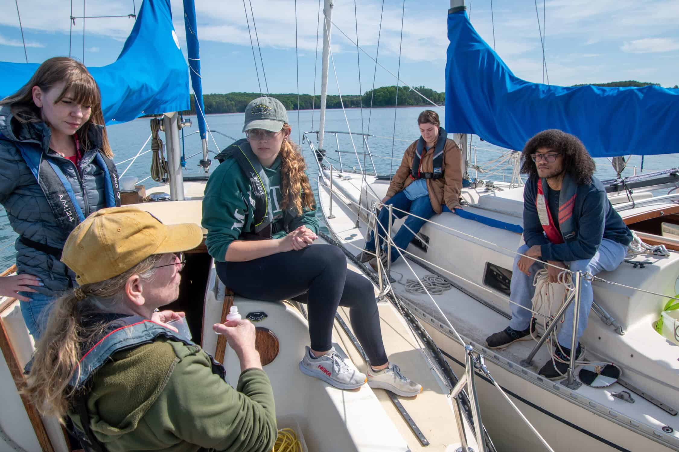 Four students on a motorboat with one lab technician wearing a light-colored ball cap in front right. Blue lake and sky and lake landscape in background.