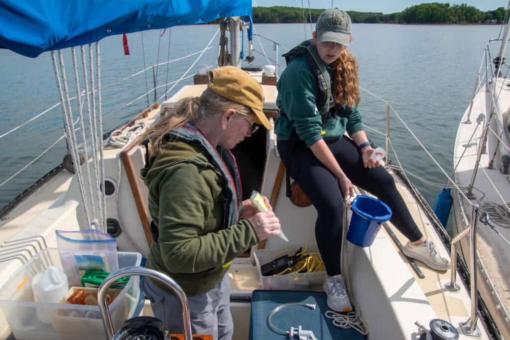 A young woman with long light hair and a ball cap on holding water quality equipment in a motorboat with an older woman on left holding and surrounded by boat and science supplies, with lake water and landscape behind them