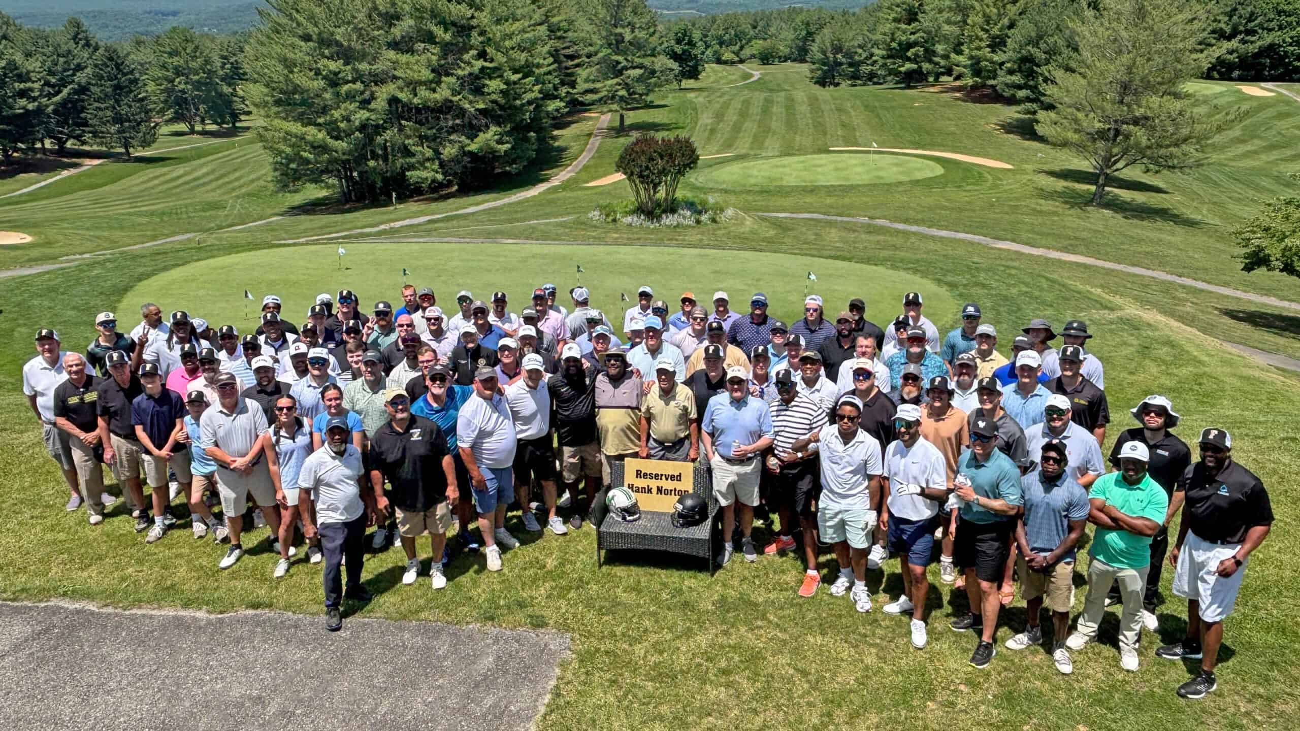 A large group of golfers pose at the edge of a golf course with trees in the background