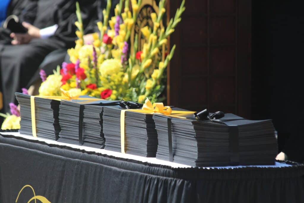 Stacks of black diplomas on table at commencement with black and gold ribbons around the stacks, and colorful tall flowers behind them.