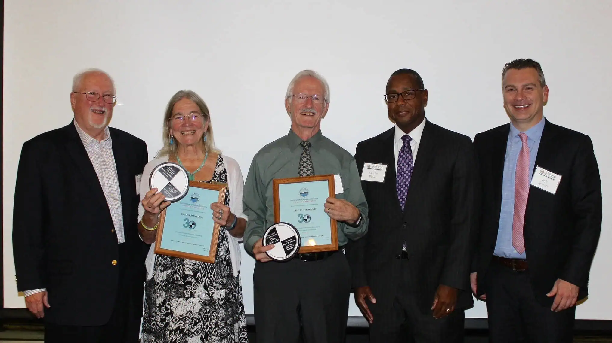 Four men in dark suits and one woman in a black and white dress holding certificates and symbols representing their work on the 30th anniversary of the Smith Mountain Lake Water Quality program