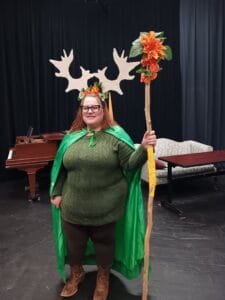 Student in a play with glasses, large antlers on her head, green clothing and cape, holding a tall staff with flowers at the top, and some low scenery and black curtain behind her.
