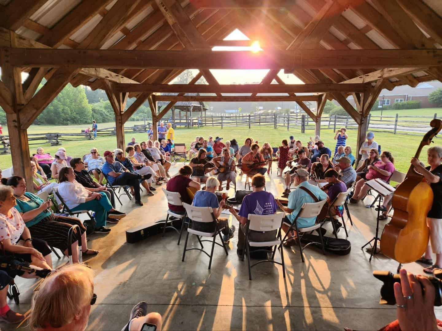 Audience gathered in an outdoor pavilion at sunset on a warm day listening to live string-band music at a Monday music jam, under roof of wooden pavilion