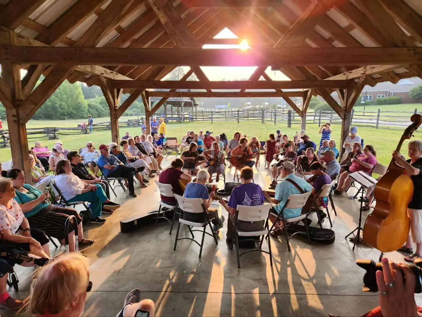 Audience gathered in an outdoor pavilion at sunset on a warm day listening to live string-band music at a Monday music jam, under roof of wooden pavilion