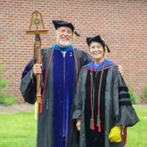 A couple of professors in graduation robes stand in front of brick building at 2019 commencement, with man holding the Ferrum College mace