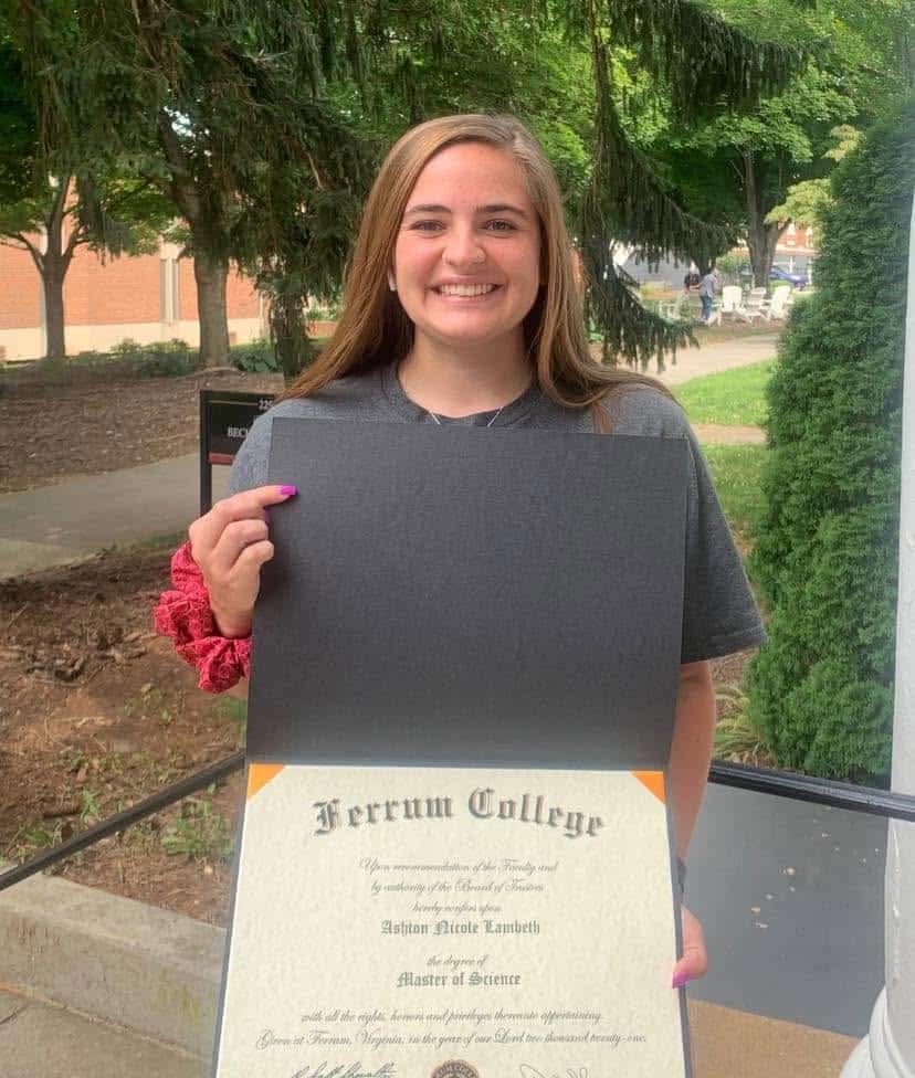 Young woman with long brown hair holding M.S. diploma open in front of her, with campus landscape in backgroun