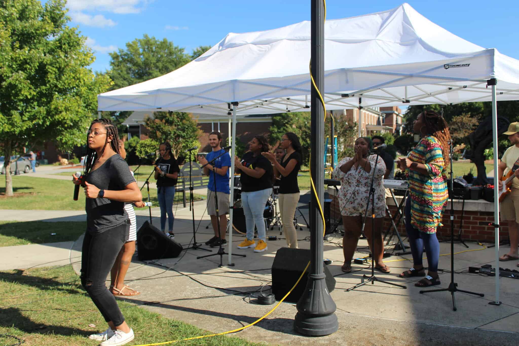 Students in casual clothes under and near a white tent performing music outdoors on campus