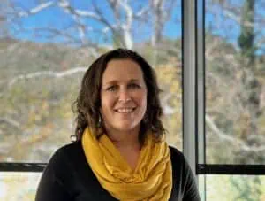 Young woman with dark hair and dress, with big gold scarf, stands in front of window