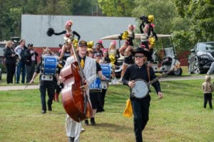Homecoming march through campus field with musicians in front carrying bass, banjo and drums, cheerleaders up high in back, in black and gold