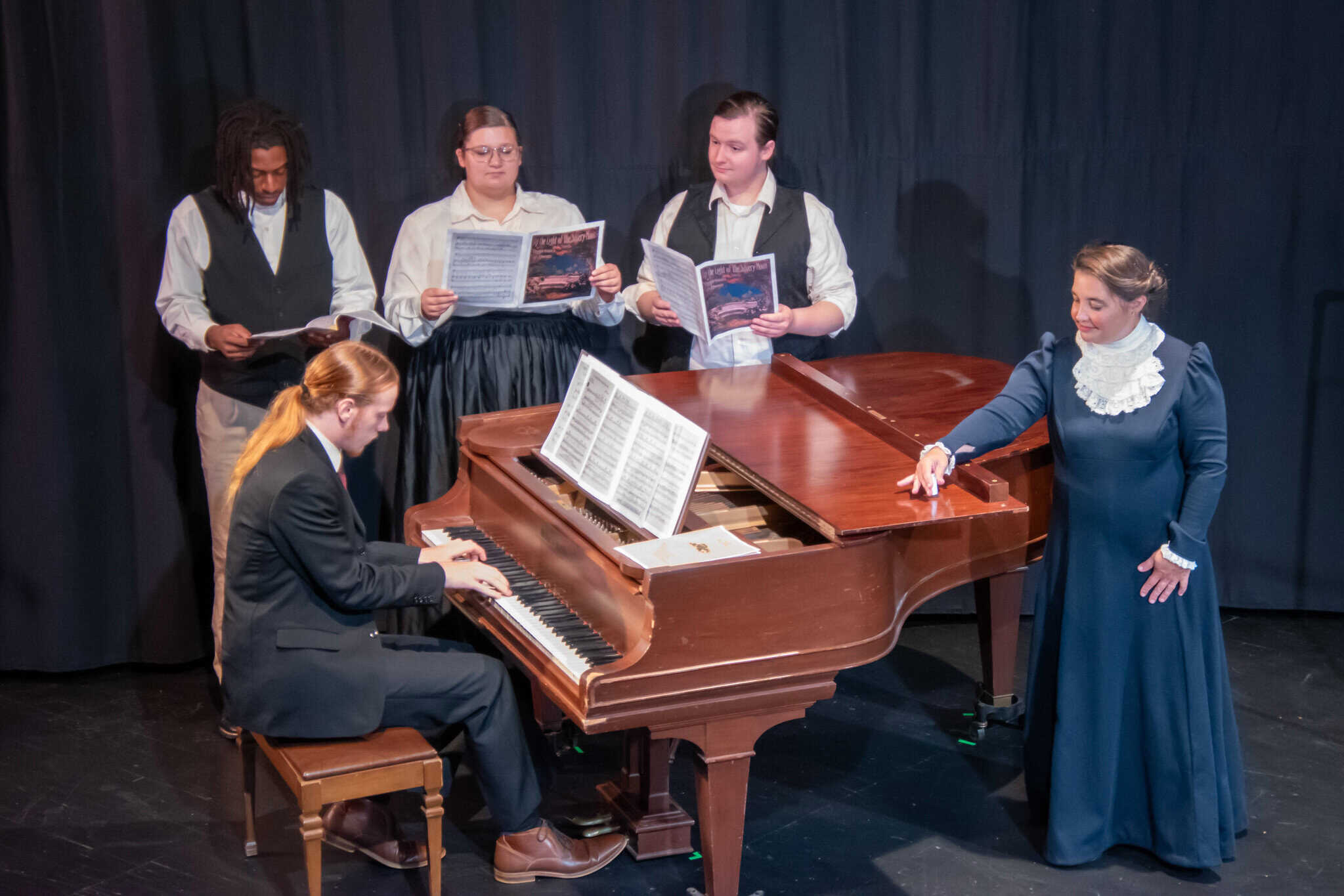 Emily Blankenship-Tucker in long navy dress at grand piano with 4 student actors singing and playing