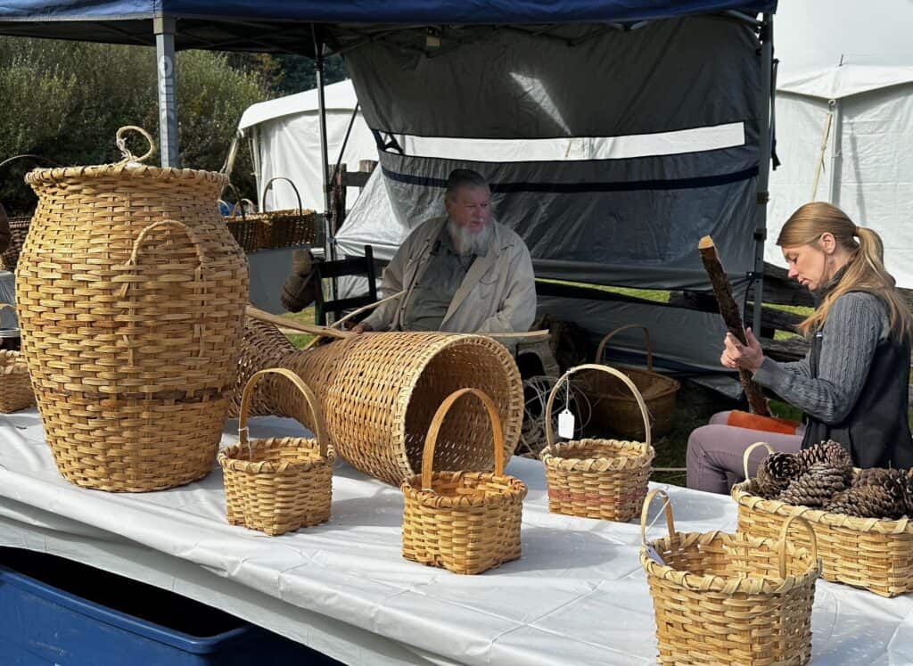 White table with different sizes of baskets on top in front of an outdoor structure at the folklife festival, with a couple people in background