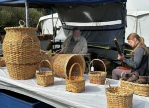White table with different sizes of baskets on top in front of an outdoor structure at the folklife festival, with a couple people in background