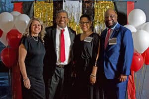 Two couples standing in front of gold, white, black, and red wall hangings, with red and white balloons beside them, one in blue suit with red striped tie, one in darker suit with black tie, women in black dresses