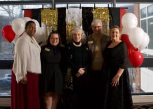 Six women standing in front of glittery gold, white, black, and red hangings, in dress outfits of black, gold, and white, with red and white balloons beside them
