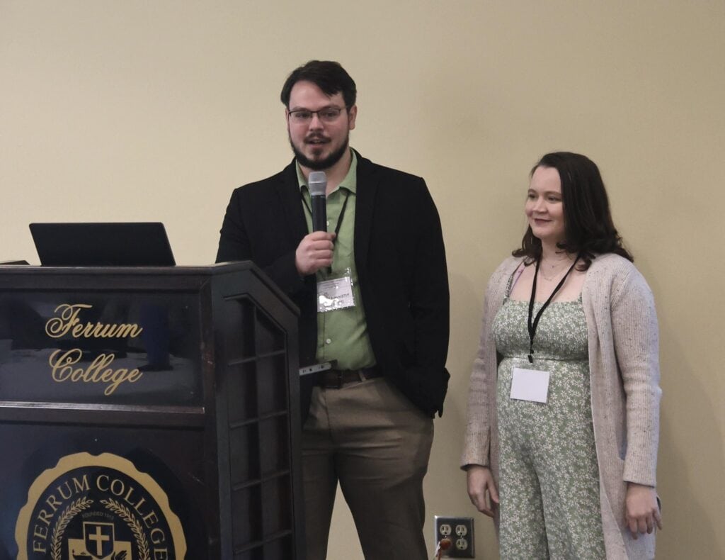 Young man with dark hair and beard, dark jacket and green shirt, holding a microphone next to a Ferrum College podium against a gold wall, with a young woman to his right, wearing a green flower-print dress and long tan sweater, with shoulder-length dark hair. Both have white name tags hanging around their necks.