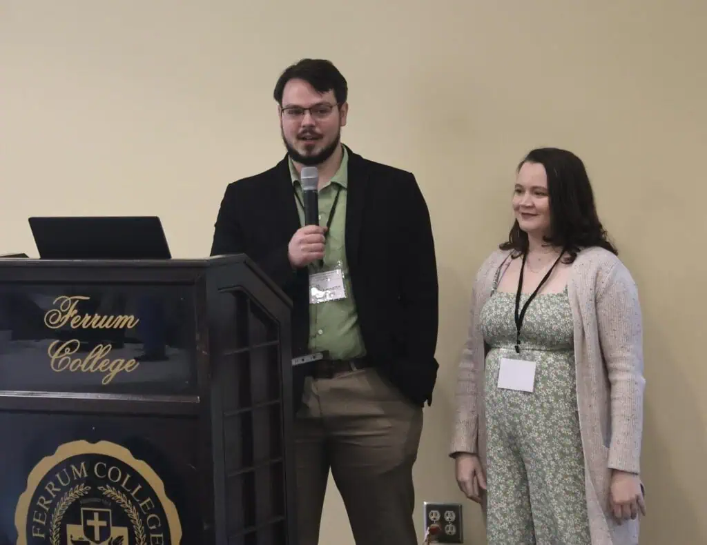 Young man with dark hair and beard, dark jacket and green shirt, holding a microphone next to a Ferrum College podium against a gold wall, with a young woman to his right, wearing a green flower-print dress and long tan sweater, with shoulder-length dark hair. Both have white name tags hanging around their necks.