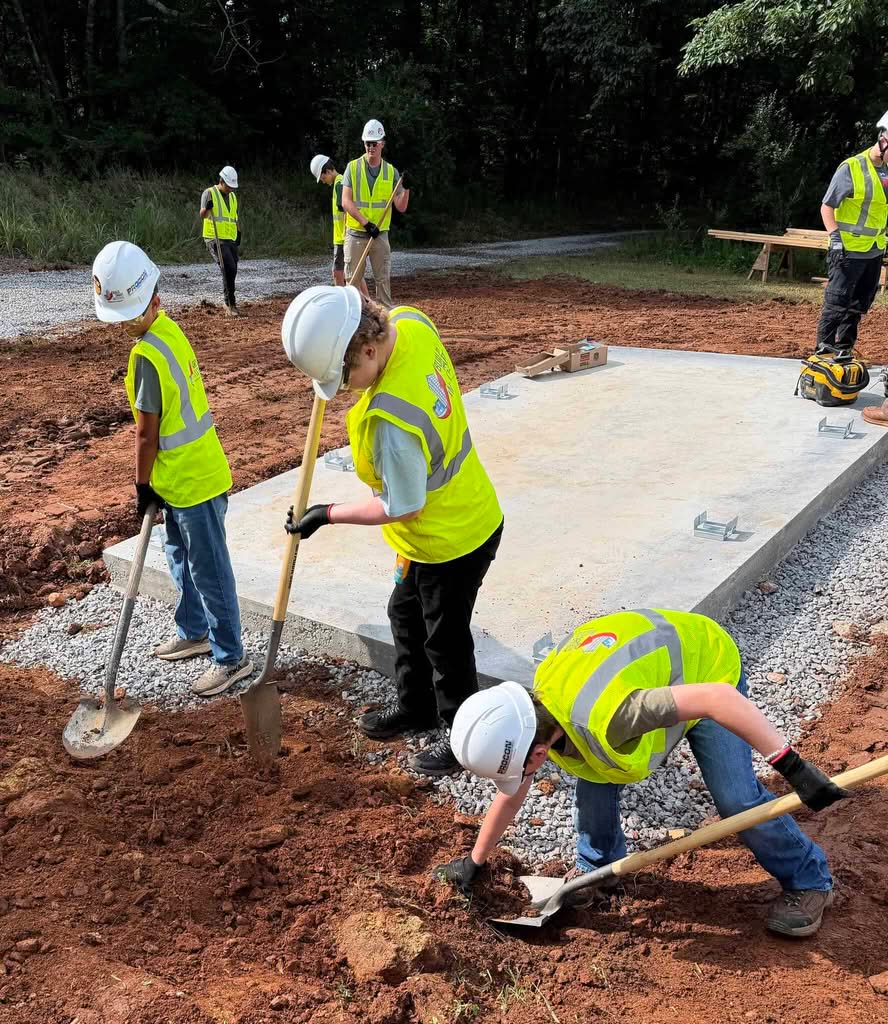 Three future builders in white helmets, neon lime vests and work gloves shoveling dirt around a new concrete floor outdoors, with several workers and trees in background.